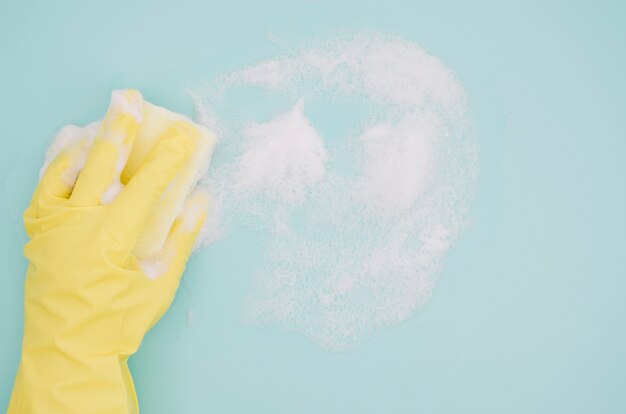 Menselijke hand die gele handschoenen draagt die blauwe achtergrond met zeep sud wassen