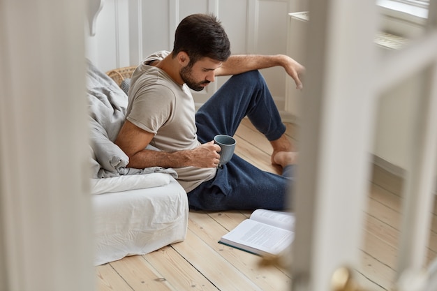 Mensen, alfabetisering, vrije tijd concept. Gerichte ongeschoren man geniet overdag van een fris drankje, gefocust in boek, leest wetenschappelijke literatuur, bereidt zich voor op het eindexamen zit op de vloer. Uitzicht vanaf deuren