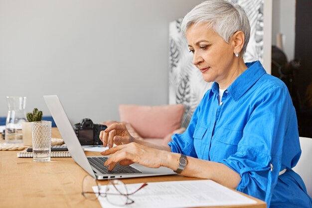 Mensen, baan, beroep, leeftijd en werk. Binnen beeld van mooie grijze haren vrouw op pensioen op zoek naar extern werk met behulp van draagbare computer. Rijpe vrouwenfotograaf die op laptop typt