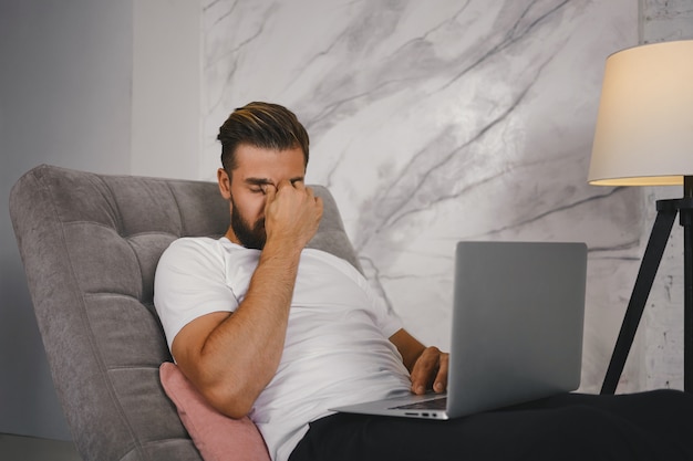 Mensen baan, overwerk en vermoeidheid concept. Foto van stijlvolle vermoeide freelancer zittend op de bank met laptop, zich uitgeput voelen tijdens het werken aan een dringend project 's avonds laat, neusbrug masseren