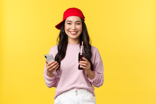 Mensen emoties, drankjes en zomer vrije tijd concept. Jonge Koreaanse tienermeisje in rode dop, berichten verzenden, smartphone gebruiken en frisdrank drinken, staande gele achtergrond tevreden.