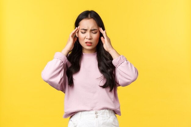 Mensen emoties, lifestyle en mode concept. Verontruste en uitgeputte aziatische vrouw sluit de ogen en raakt de slapen aan, heeft migraine, voelt hoofdpijn of duizelige, gele achtergrond.