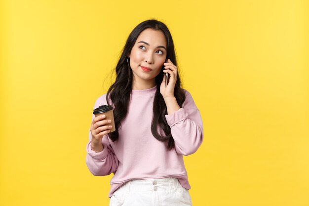 Mensen emoties, lifestyle vrije tijd en schoonheid concept. Leuke stijlvolle aziatische vrouw die attent opkijkt terwijl ze op mobiele telefoon praat en koffie drinkt uit een afhaalbeker, gele achtergrond.