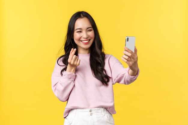 Mensen emoties, lifestyle vrije tijd en schoonheid concept. Vrolijk Aziatisch meisje over gele achtergrond die selfie op mobiele telefoon neemt, app voor fotofilters gebruikt en hartgebaar toont.