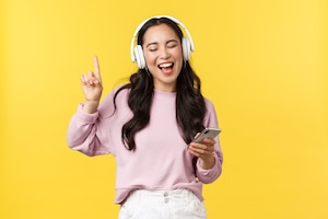 Mensen emoties, lifestyle vrije tijd en schoonheid concept. zorgeloze gelukkige aziatische vrouw die muziek luistert in draadloze koptelefoons, mobiele telefoon vasthoudt, meezingt met favoriete liedje, gele achtergrond.