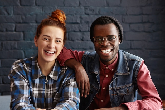 Gratis foto mensen en levensstijl concept. aantrekkelijke jonge man met bril en stijlvolle hipster zwarte hoed zijn elleboog leunend op de schouder van zijn mooie roodharige vriendin tijdens bijeenkomst in coffeeshop