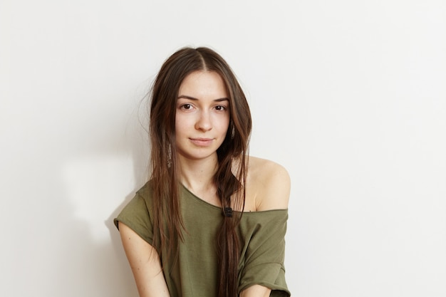 Mensen en levensstijl concept. Headshot van mooie blanke brunette tiener vrouw met wild kapsel dragen trendy legergroen t-shirt met een schouder opdagen, poseren geïsoleerd op witte muur