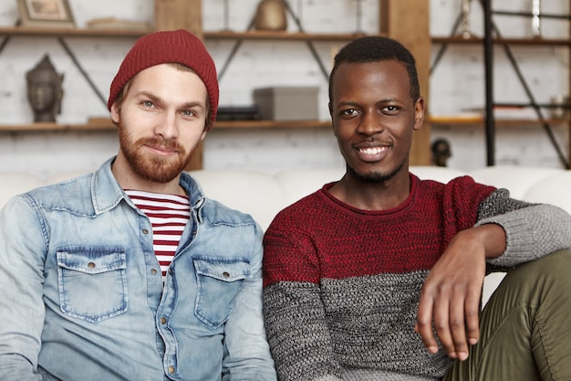 Gratis foto mensen en levensstijl concept. twee gelukkige jonge mannen van verschillende etnische groepen samen tijd doorbrengen, zittend op de bank dicht bij elkaar. stijlvolle witte man in hoed binnenshuis rusten met zijn zwarte vriend