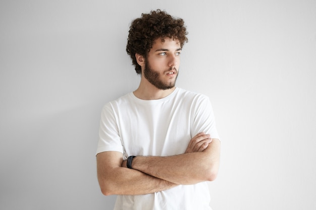 Mensen en levensstijlconcept. Portret van modieuze jonge Kaukasische bebaarde hipster die witte t-shirt draagt die met gevouwen wapens stelt, wegkijkend met ernstige bedachtzame uitdrukking