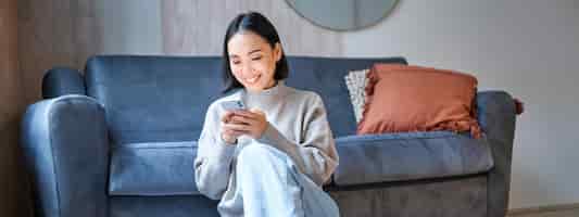 Gratis foto mensen en technologie lachende aziatische vrouw om thuis te zitten met behulp van haar mobiele telefoon bericht te typen