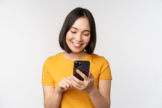 Mensen en technologieconcept Glimlachend Aziatisch meisje dat smartphone sms't op mobiele telefoon die zich tegen witte achtergrond bevindt
