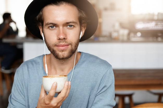 Gratis foto mensen en vrije tijd concept. headshot van aantrekkelijke jonge hipster in trendy zwarte hoed koffie drinken en muziek luisteren