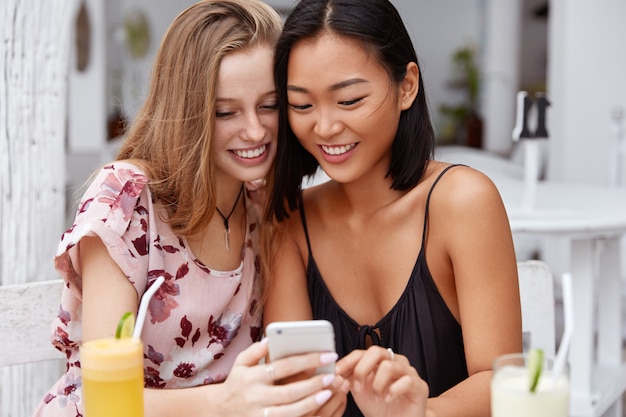 Mensen etniciteit, technologie, entertainment. Tevreden Aziatische en Europese vrouwen recreëren samen in een coffeeshop, kijken positief naar de smartphone