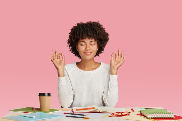 Mensen, harmonie en werkconcept. Tevreden vrouw met donkere huid en Afro-kapsel, mediteert op het werk, houdt de ogen gesloten