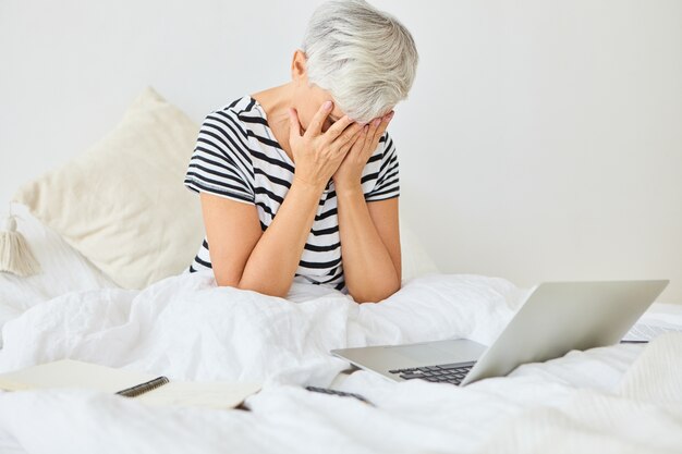 Mensen, leeftijd en technologie. Senior vrouw zittend op bed met depressieve blik, gezicht met beide handen bedekkend alsof ze huilde, gestrest omdat ze per ongeluk alle gezinscomputers op laptop heeft verwijderd