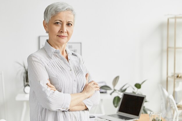 Mensen, leeftijd, technologie en baanconcept. Knappe serieuze vrouwelijke stafmedewerker van middelbare leeftijd met kort pixiekapsel dat zich op kantoor bevindt met gekruiste armen op de borst, haar houding die vertrouwen uitdrukt