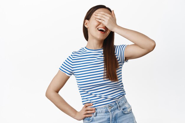 Mensen levensstijl. Portret van een jonge gelukkige vrouw van 20 jaar, die ogen bedekt met de hand, wachtend op verrassing, verstoppertje, glimlachend opgewonden, staande tegen een witte achtergrond