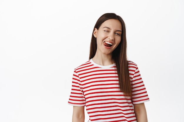 Mensen levensstijl. Portret van mooi meisje knipogen en tong positieve, blije gezichtsuitdrukking van jonge vrouw in casual t-shirt, witte achtergrond tonen. Ruimte kopiëren