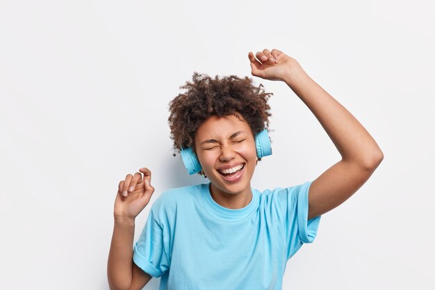 Mensen levensstijl vrije tijd concept. Energieke vrolijke Afro-Amerikaanse vrouw met krullend haar danst zorgeloos steekt armen op voelt zich geamuseerd geniet van favoriete muziek draagt draadloze koptelefoon blauw t-shirt