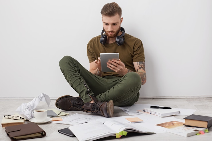 Gratis Foto mensen, moderne technologie en onderwijsconcept. stijlvolle man met baard draagt laarzen, zit gekruiste benen op de vloer,
