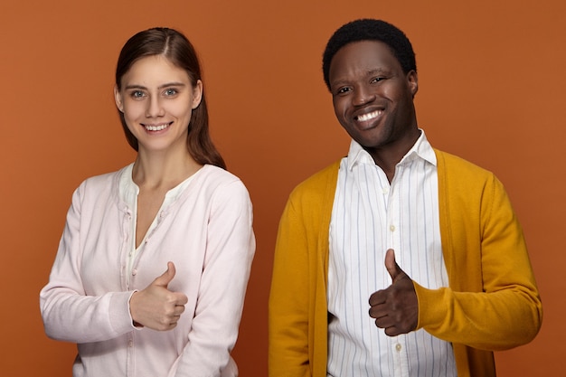 Mensen, succes, tekens en gebarenconcept. Succesvolle vriendelijk ogende zelfverzekerde jonge zwarte mannelijke en blanke vrouw, duimen opdagen gebaar, genieten van samenwerken in team, gelukkig lachend