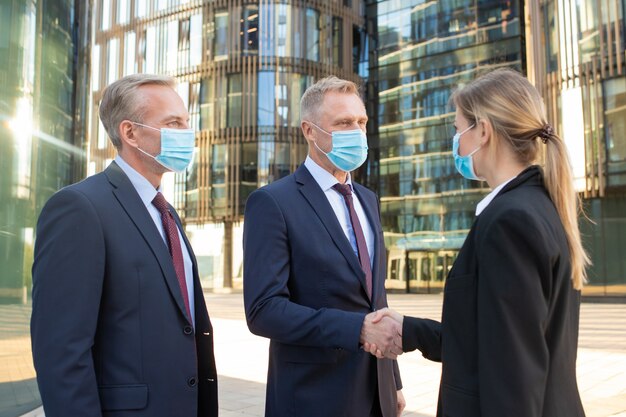 Mensen uit het bedrijfsleven dragen gezichtsmaskers, staan in de buurt van kantoorgebouwen, schudden elkaar de hand, ontmoeten elkaar en praten in de stad. Zijaanzicht, lage hoek. Bedrijf tijdens uitbraak concept