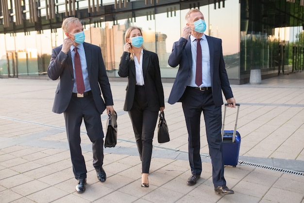 Mensen uit het bedrijfsleven passen zich aan of zijn klaar om gezichtsmaskers af te doen tijdens het wandelen met bagage buitenshuis, in de buurt van kantoorgebouwen. Zakenreis en einde van epidemisch concept