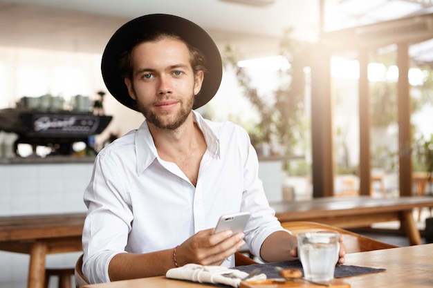 Mensen, vrije tijd en moderne technologie. Jonge student die met gelukkige blik van hoge snelheidsinternetverbinding op zijn slimme telefoon geniet. Modieuze man in trendy hoofddeksels met behulp van elektronische apparaat in café