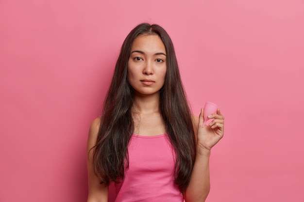 Menstruele bescherming, gynaecologie en vrouwelijke hygiëne concept. Ernstige brunette etnische vrouw houdt menstruatiecup, gebruikt geen afvalabsorberend product in de bloedperiode, geïsoleerd op roze muur