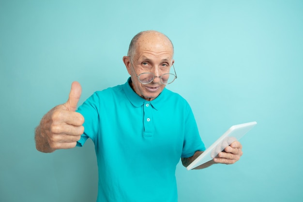 Met behulp van de tablet, duim omhoog. Kaukasische senior man portret op blauwe studio achtergrond. Mooi mannelijk emotioneel model. Concept van menselijke emoties, gezichtsuitdrukking, verkoop, welzijn, advertentie. Copyspace.