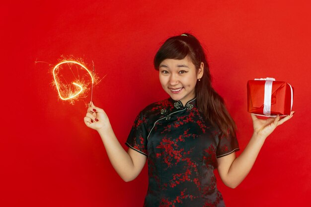 Met geschenkdoos en helder sterretje. Gelukkig Chinees nieuwjaar. Aziatisch jong meisje portret op rode achtergrond. Vrouwelijk model in traditionele kleding ziet er gelukkig uit. Copyspace.