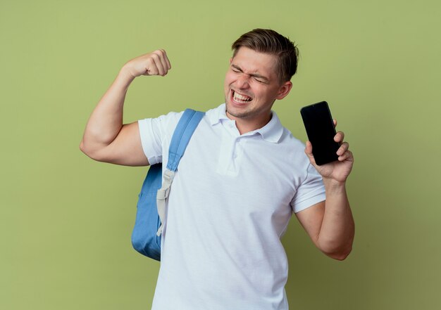 Met gesloten ogen blije jonge knappe mannelijke student die achterzak draagt die telefoon houdt en ja gebaar toont dat op olijfgroen wordt geïsoleerd