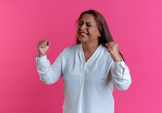 Met gesloten ogen blije ongedwongen blanke vrouw van middelbare leeftijd die ja gebaar toont dat op roze muur wordt geïsoleerd