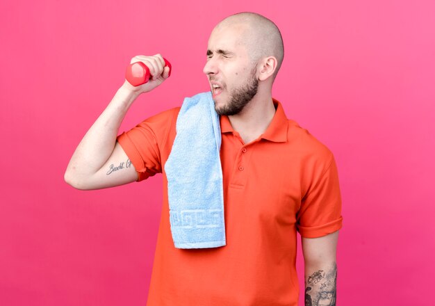 Met gesloten ogen gespannen sportieve jongeman trainen met halter met handdoek op schouder geïsoleerd op roze muur