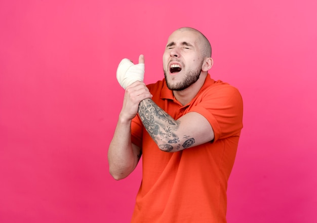 Met gesloten ogen gewonde jonge sportieve man met pols verband greep hand geïsoleerd op roze muur