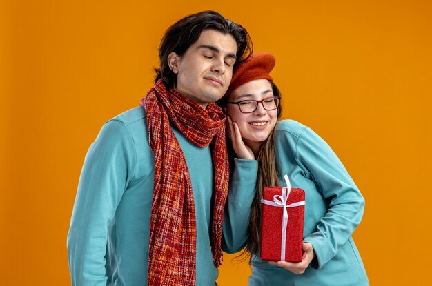 Met gesloten ogen jong koppel op Valentijnsdag man met sjaal meisje met geschenkdoos geïsoleerd op oranje achtergrond