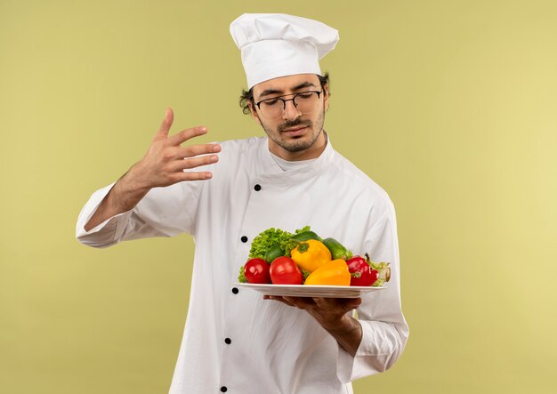 Met gesloten ogen jonge mannelijke kok die eenvormige chef-kok en glazen dragen die en groenten op plaat houden die op groene muur wordt geïsoleerd