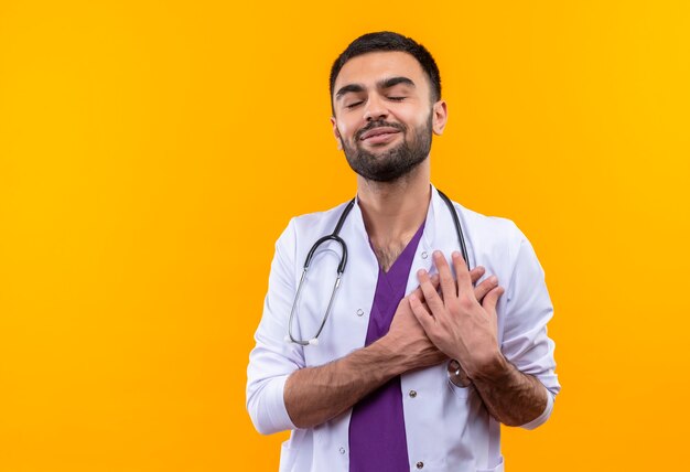 Met gesloten ogen legde de jonge mannelijke arts die stethoscoop medische toga draagt zijn handen op hart op geïsoleerde gele muur