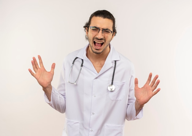 Met gesloten ogen spreidt de boze jonge mannelijke arts met optische bril die een wit gewaad met een stethoscoop draagt zijn handen uit op geïsoleerde witte muur met exemplaarruimte