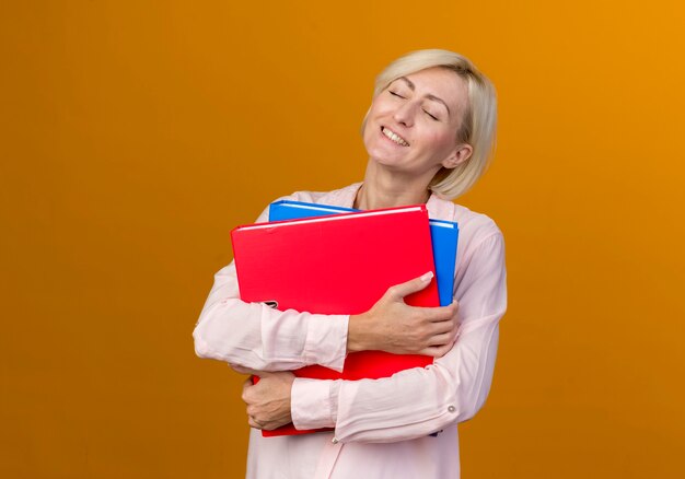 Met gesloten ogen tevreden jonge blonde Slavische vrouw met mappen geïsoleerd op oranje muur