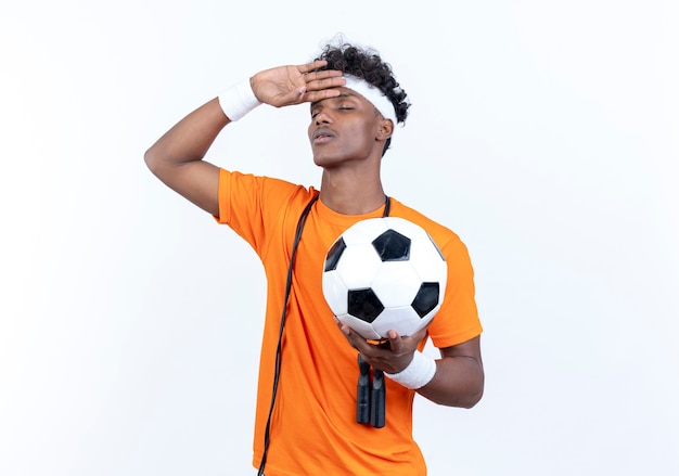 Met gesloten ogen vermoeide jonge Afro-Amerikaanse sportieve man met hoofdband en polsband die bal vasthoudt en hand op het voorhoofd legt met springtouw op schouder