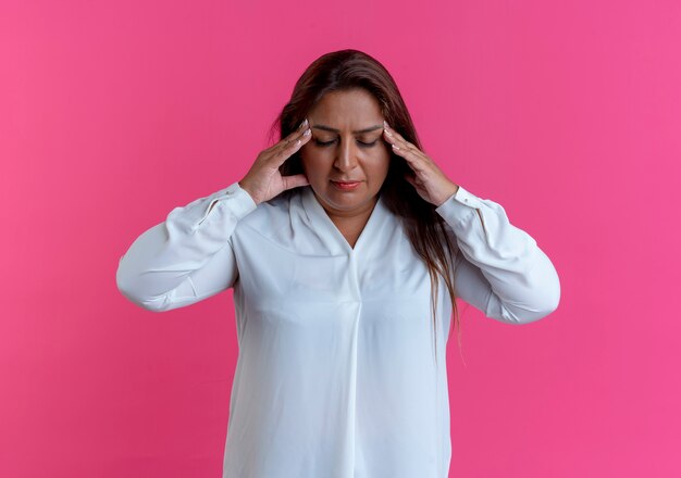 Met gesloten ogen vermoeide toevallige Kaukasische vrouw van middelbare leeftijd die handen op voorhoofd zetten dat op roze muur wordt geïsoleerd