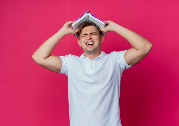 Met gesloten ogen vreugdevolle jonge knappe mannelijke student bedekt hoofd met boek geïsoleerd op roze muur