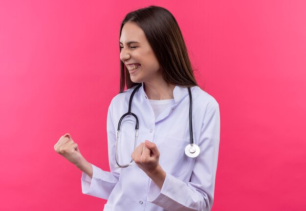 Met gesloten ogen vrolijk jong artsenmeisje die stethoscoop medische toga dragen die ja gebaar op geïsoleerde roze muur tonen