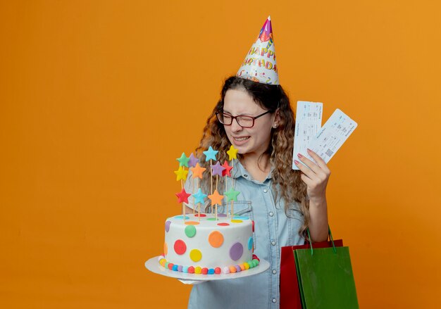 Met gesloten ogen vrolijk jong meisje bril en verjaardag GLB bedrijf kaartjes en verjaardagstaart met cadeau tassen geïsoleerd op een oranje achtergrond