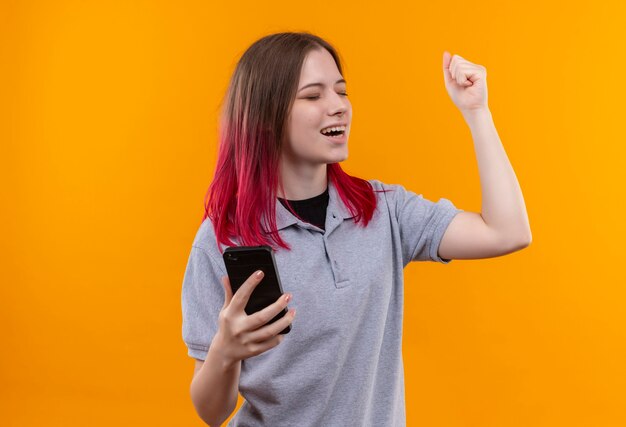 Met gesloten ogen vrolijk jong mooi meisje die de grijze telefoon van de t-shirtholding dragen die ja gebaar op geïsoleerde gele muur toont