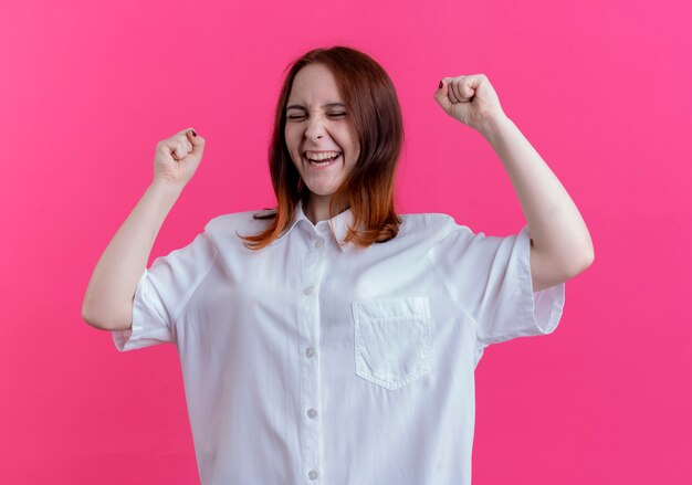 Met gesloten ogen vrolijk jong roodharigemeisje die vuisten opheffen die op roze worden geïsoleerd