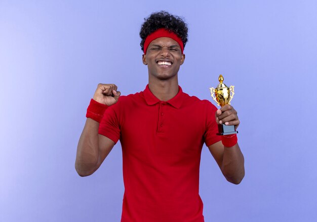 Met gesloten ogen vrolijke jonge afro-amerikaanse sportieve man met hoofdband en polsbandje winnaar beker houden en ja gebaar geïsoleerd op blauw tonen