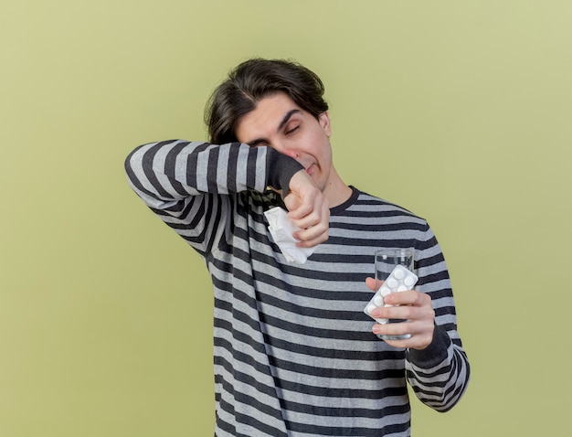 Met gesloten ogen zwakke jonge zieke man met glas water met pillen en afvegen gezicht met arm geïsoleerd op olijfgroene achtergrond