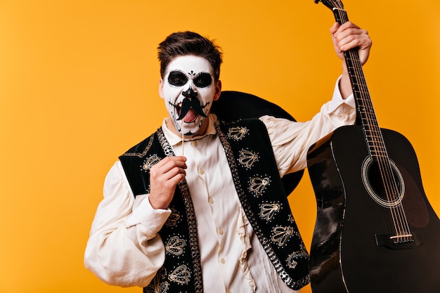 Mexicaanse bruinogige man met gezichtskunst in vorm van schedel schreeuwt emotioneel, poseren met valse snorren en gitaar in zijn handen op oranje muur.